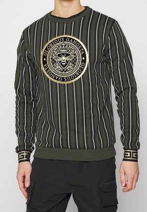 Mand iført grøn og sort vertikalt striped sweater med "Glorious Gangsta" guldemblem med en bi og krone på brystet.