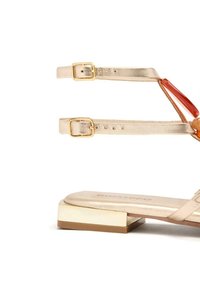 Las sandalias metálicas doradas cuentan con dos delgadas correas en el tobillo y un tacón cuadrado con un acento crema. La plantilla tiene la marca "Gioseppo".