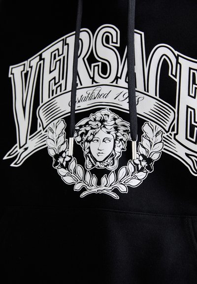 Felpa nera con testo bianco "VERSACE", logo della testa di Medusa circondato da allori e lacci neri con punte argentate.