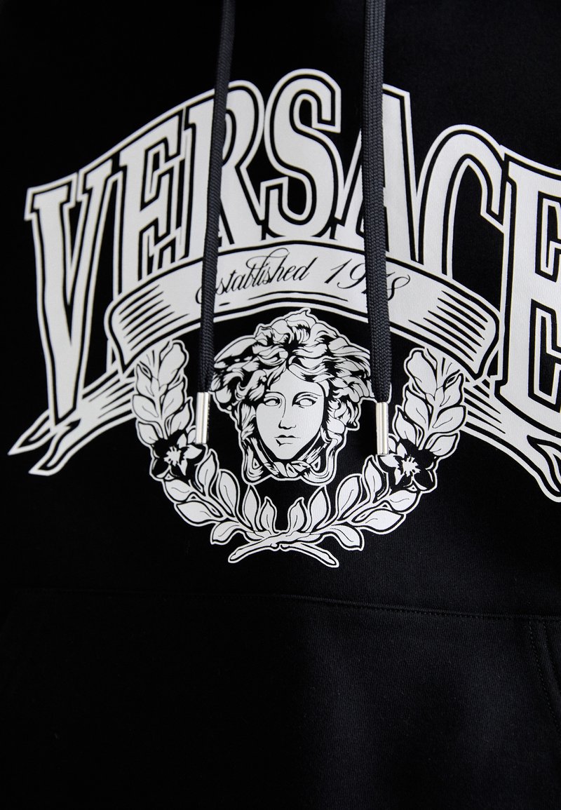 Felpa nera con testo bianco "VERSACE", logo della testa di Medusa circondato da allori e lacci neri con punte argentate.