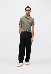 Tricou polo verde măsliniu cu un mic logo brodat, asortat cu pantaloni largi negri și adidași albi cu talpă bej.