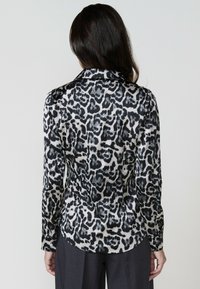 Una donna con capelli lunghi indossa una blusa a stampa leopardata, vista da dietro, abbinata a pantaloni scuri; sfondo minimale.