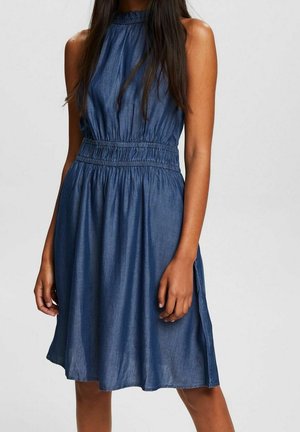 Kobieta w niebieskiej sukience chambray bez rękawów z marszczonym wysokim dekoltem i elastycznym paskiem w talii, stojąca na jednolitym tle.
