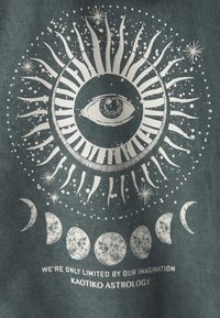 Tejido verde con un gráfico celestial: un ojo en el centro, rodeado de rayos de sol, estrellas y fases de la luna, con texto debajo.