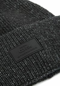 Zrobiona na drutach czarna czapka beanie z teksturowanym wzorem, z czarną prostokątną metką wytłoczoną z tekstem i współrzędnymi.