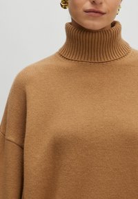 Kamel-farbener Rollkragenpullover aus geripptem Strickstoff, mit hohem Hals und lockerer Passform. Weiche Textur mit dezenten Nähten.