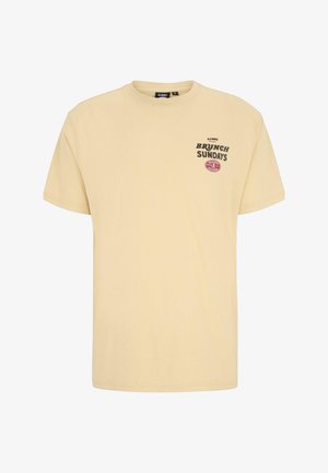Béžové tričko s krátkým rukávem a černým textem "Brunch Sundays" a malým červeným oválným logem na levé hrudi, kulatý výstřih.
