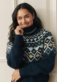 Adīts džemperis ar augstu apkakli, tumši zilu pamatu un ģeometriskiem rakstiem zaļā, dzeltenā un baltā krāsā. Faktūrēts audums ar riblotām aprocēm.