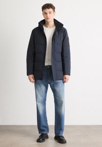 Giacca puffer blu navy con cappuccio staccabile, chiusura zip, tasche laterali e tessuto testurizzato. Abbinata a jeans azzurri chiari e scarpe nere.