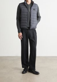 Szara puchowa kamizelka z wysokim kołnierzem, zapinana na zamek błyskawiczny, z naszywką z logo, noszona na ciemnej koszuli z długim rękawem i czarnych spodniach, do tego czarne buty.