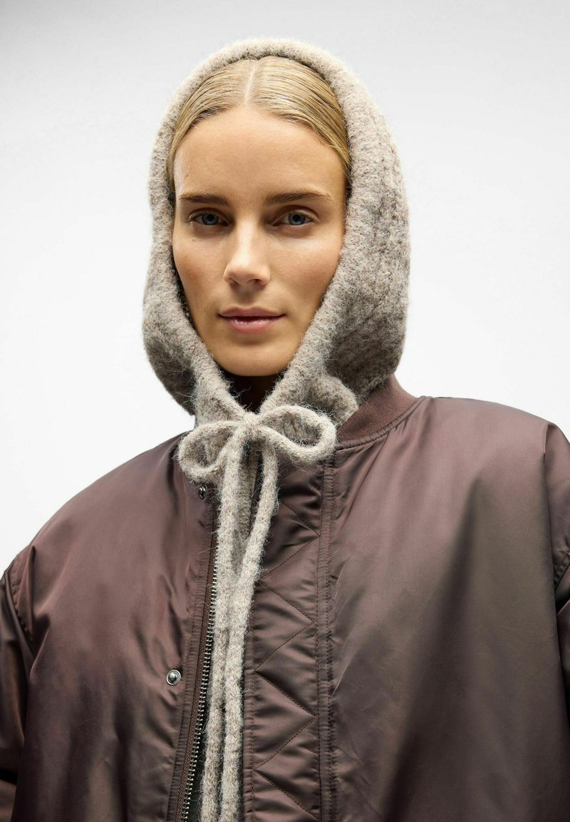 Veste aviateur marron avec fermeture éclair, accompagnée d'une capuche en tricot gris clair ornée d'un nœud papillon. Texture lisse avec une apparence confortable.