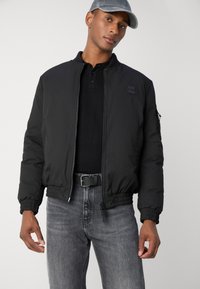 Svart bomberjakke med glidelås foran, ribbestrikkede mansjetter og linning. Båret over en svart polo-skjorte og kombinert med grå jeans og en grå caps.