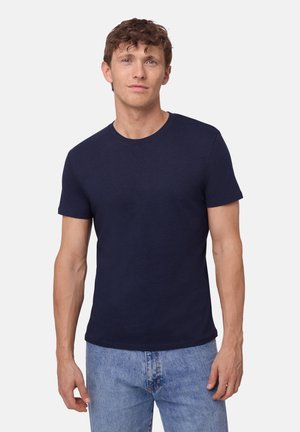 Homme aux cheveux bruns courts et bouclés portant un t-shirt bleu marine uni à manches courtes et un jean bleu clair, sur un fond blanc.