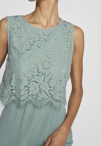 Un haut en dentelle vert menthe clair avec un ourlet festonné, des motifs floraux et un col rond. Le matériau présente une finition délicate et texturée.