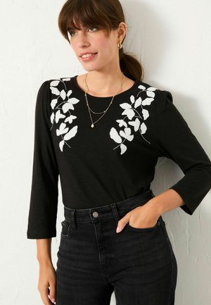Schwarzes Langarmoberteil mit weißer floralem Stickerei an den Schultern, rundem Halsausschnitt und dezenten Puffärmeln, kombiniert mit hochgeschnittenen dunklen Jeans.