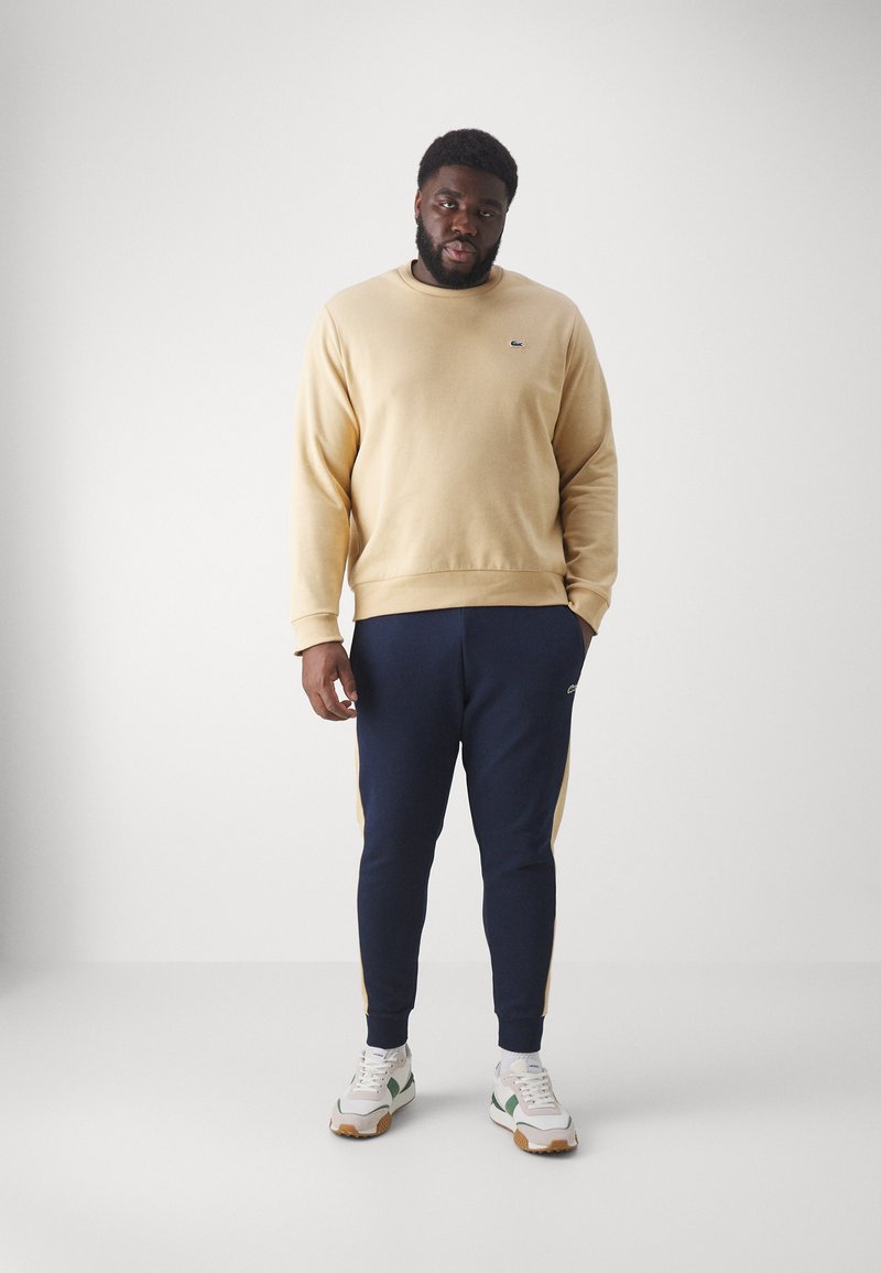 Hellgelbes Sweatshirt mit gerippten Bündchen, kombiniert mit marineblauen Jogginghosen mit seitlichem Streifen. Weiße Sneakers mit grünen und braunen Akzenten.