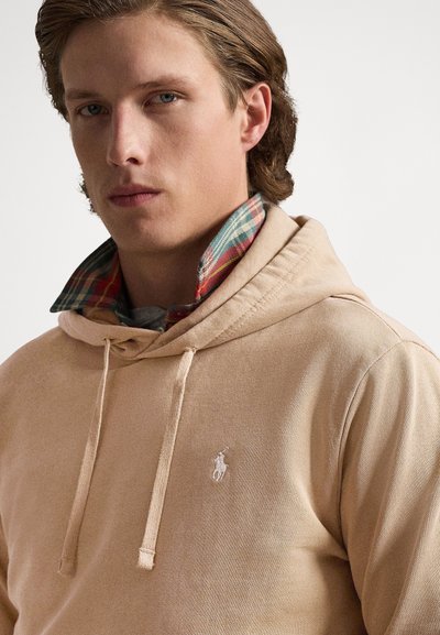 Sweatshirt bege com capuz feita de tecido macio com uma textura subtil, apresentando um bolso frontal em estilo canguru e um pequeno logótipo bordado no peito.