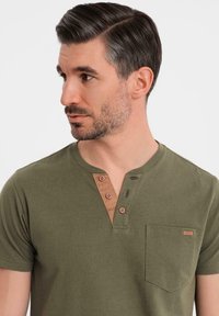 Oliwkowa koszulka z krótkim rękawem z brązowym kołnierzykiem henley, z trzema guzikami i kieszenią na piersi z małą etykietką. Miękki materiał.