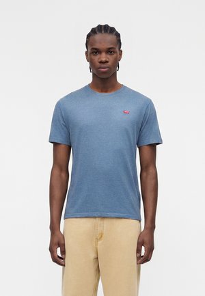 Νέος άνδρας με πλεξούδες και μπλε μπλούζα Levi's και μπεζ παντελόνι, στέκεται μπροστά σε απλό φωτεινό φόντο.