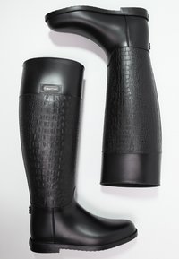 Botas negras de caña alta con un upper liso y un patrón texturizado de cocodrilo en la mitad inferior. Presentan un logo de marca y una suela plana.