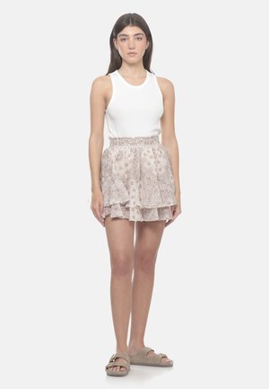 Donna in piedi che indossa una canotta bianca senza maniche, gonna beige a balze con motivo e sandali beige open-toe con cinturini a fibbia su sfondo bianco.