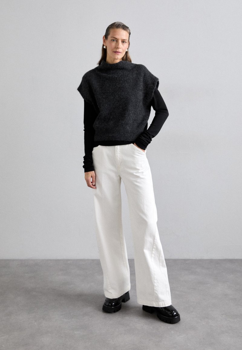 Top oversize senza maniche in maglia nera sovrapposto a una camicia nera a maniche lunghe, abbinato a pantaloni bianchi a gamba larga e scarpe chunky nere.