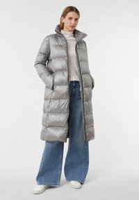 Lange zilveren pufferjas met een hoge kraag, ritsluiting en zijzakken. Gemaakt over een lichte trui en wijde jeans. Gladde, glanzende textuur.