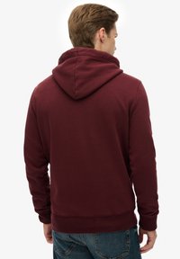 Sudadera con capucha de color burdeos con textura suave, bolsillo tipo canguro y puños acanalados. Vista trasera, visibles unos vaqueros de mezclilla azules en contraste.