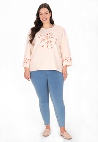 Hanorac roz deschis cu un model floral, mâneci lungi și detaliu brodat cu floare în centru. Asortat cu blugi skinny albaștri și pantofi bej.