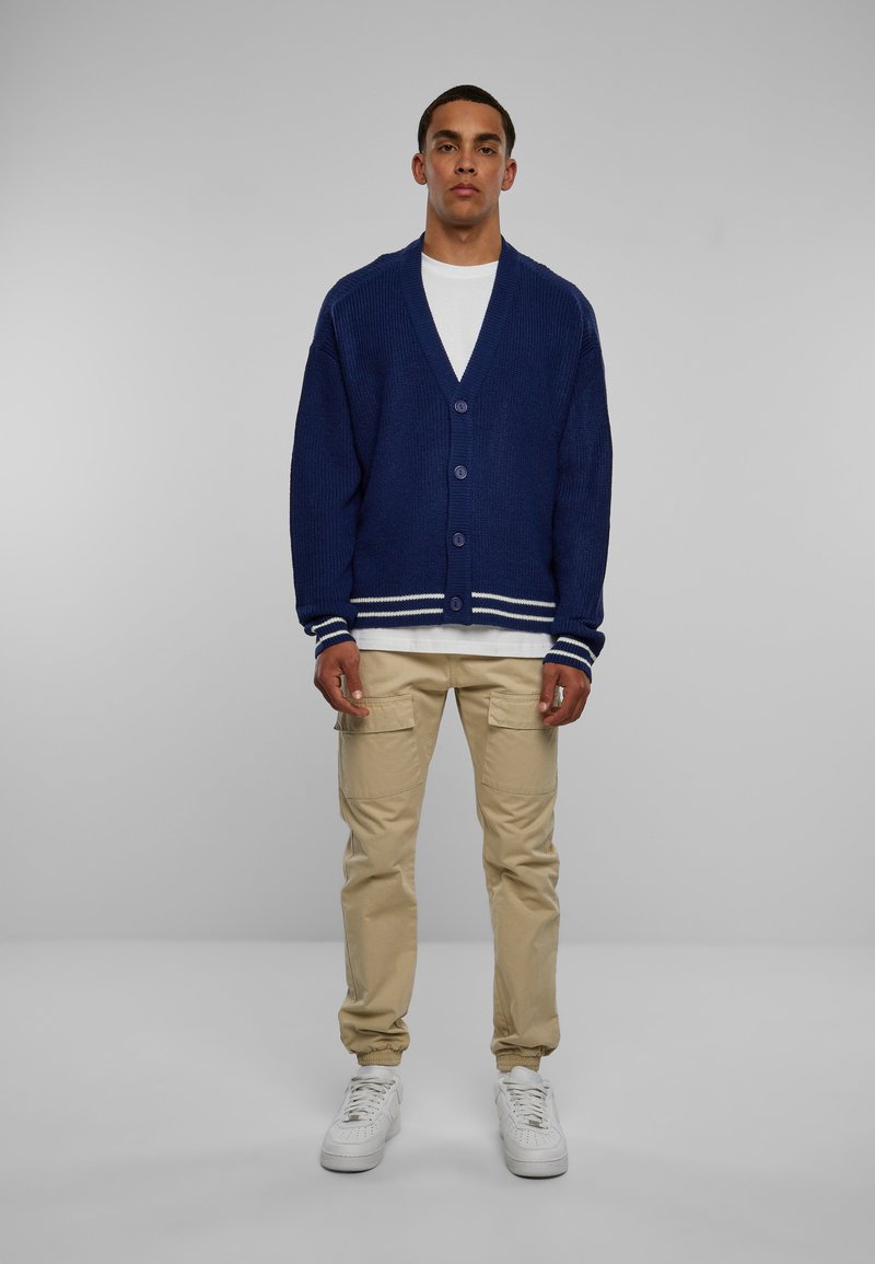 Mørkeblå strikket cardigan med V-hals, fire knapper og hvite striper på ermene, kombinert med beige cargo-bukser og hvite sneakers.