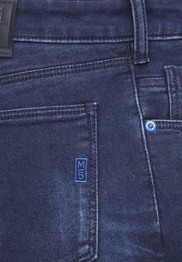 Jean en denim bleu foncé avec une étiquette logo M5, coupe droite, et poche avant avec des coutures bleues et un bouton en métal.