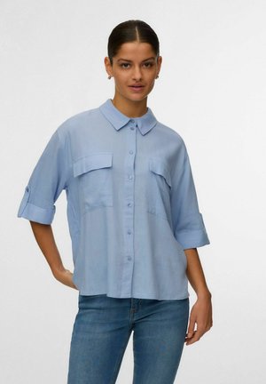 Žena s tmavými vlasy svázanými do nízkého drdolu, oblečená v světle modré košili s rozhalenými rukávy a modrými džínami, stojící před jednobarevným pozadím.