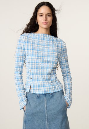 Vrouw draagt een blauw-wit geruit topje met lange mouwen en zij knopen en een lichtblauwe denim rok met handen in de zakken.