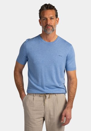 Man met baard die een lichtblauw gebreid shirt met korte mouwen en beige broek draagt, staand met één hand in zijn zak tegen een effen achtergrond.