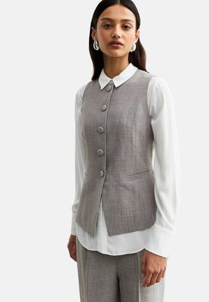 Gilet grigio su misura con sei bottoni, colletto marcato e due tasche, indossato sopra una camicia bianca a maniche lunghe con dettagli ai polsini.