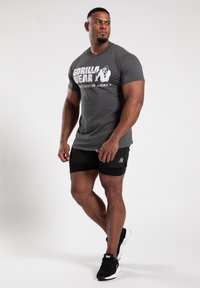 Grau T-Shirt mit dem Aufdruck "GORILLA WEAR UNITED STATES OF AMERICA"; kombiniert mit schwarzen Shorts und schwarzen Sportschuhen, posierend vor einem weißen Hintergrund.