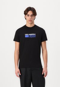 Juodas medvilnės marškinėliai su baltu ir mėlynu "KARL LAGERFELD JEANS" logotipu priekyje. Klasikinis apvalus kaklas ir trumpomis rankovėmis.