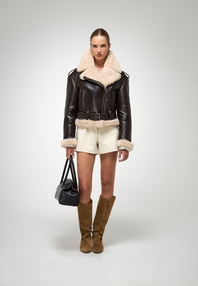 Bruine leren cropped jack met beige schapenvacht kraag en manchetten, gecombineerd met lichte shorts en bruine suède laarzen tot de knie, terwijl ze een zwarte handtas vasthoudt.