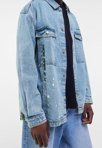 Ljusblå denimjacka med knäpps i midjan, två bröstfickor, stänkdetaljer och textgrafik på vänster ärm.