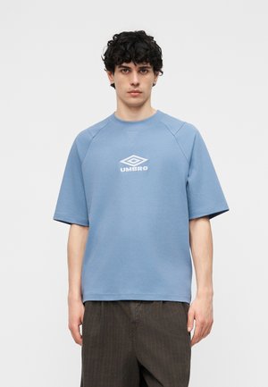 Νέος άντρας με σκούρα σγουρά μαλλιά, ντυμένος με ανοιχτό μπλε φανελάκι με κοντά μανίκια Umbro και σκούρα καφέ παντελόνια, μπροστά σε απλό φόντο.