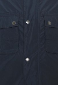 Navy blue padded jacket with metallic snap buttons, two chest pockets, and a smooth texture. Features a zip closure concealed by a flap.