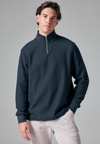 Dunkelblauer Halb-Reißverschluss-Pullover aus strukturiertem Stoff; mit hohem Kragen und langen Ärmeln. Getragen mit hellen Hosen.