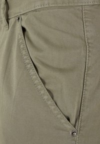 Khaki-housut, joissa on sileä, teksturoitu kangas, sivussa läpällinen tasku ja metallinen neppariyksityiskohta.