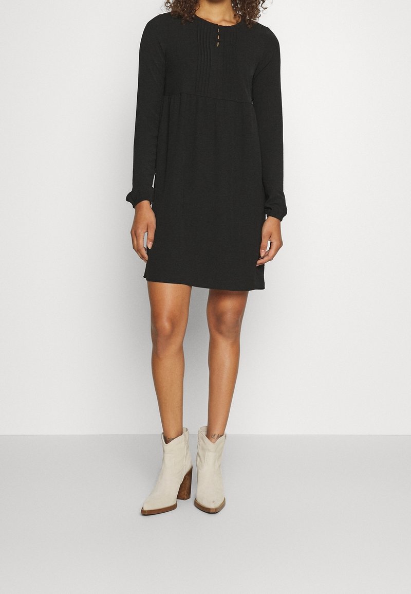 Femme portant une robe noire à manches longues jusqu'aux genoux et des bottines à talons blanc cassé, debout devant un fond blanc uni.