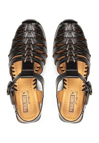 Sandalias de cuero negro con un diseño trenzado, interior en tono marrón claro y correa ajustable con un hebilla circular. Suela plana con detalles de costura visibles.