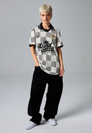 Korte mouwen, geruite shirt met zwarte kraag; bevat 'Land Back' grafisch ontwerp en patchdetails; gecombineerd met losse zwarte broek en witte schoenen.