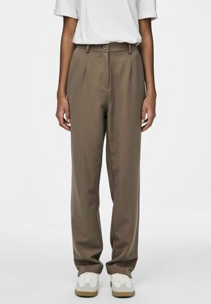 Pantalones de traje marrones con cierre de botón, pliegue sencillo y bolsillos laterales, combinados con zapatillas blancas sobre un fondo liso.