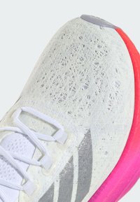 Bílé sportovní tenisky s texturovaným svrškem, stříbrnými akcenty a jasně růžovou podrážkou. Obsahují šňůrky a vrstvené vzory na materiálu.