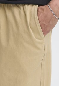 Pantalon en coton beige à coupe décontractée, doté de poches latérales, d'une texture subtile et d'une taille froncée. Design simple sans motifs.