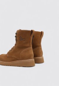 Botas de tobillo de ante marrón con punta redondeada, cordones en la parte delantera y cremallera lateral. Suela de goma gruesa y detalle del logo grabado en el caña.